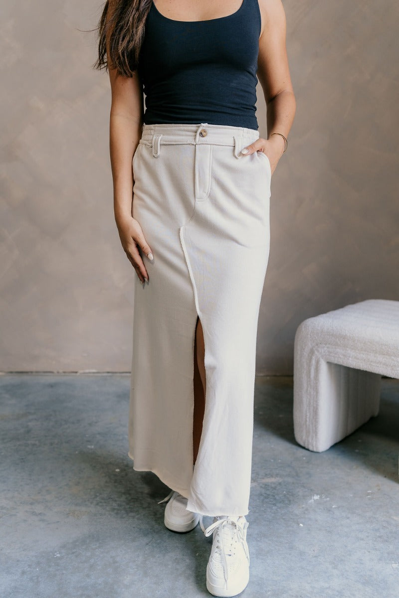 Front view of model wearing the Tori Cream Front Slit Midi Skirt which features cream fabric, a front slit, a tortoise button closure, a back elastic waistband, belt loops, front pockets, distressed details in the back, and a raw hem.
