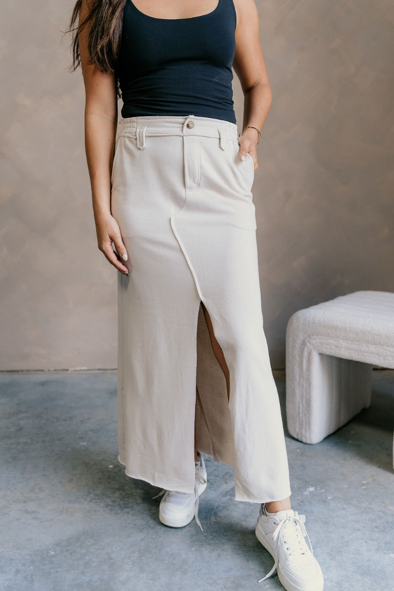 Front view of model wearing the Tori Cream Front Slit Midi Skirt which features cream fabric, a front slit, a tortoise button closure, a back elastic waistband, belt loops, front pockets, distressed details in the back, and a raw hem.