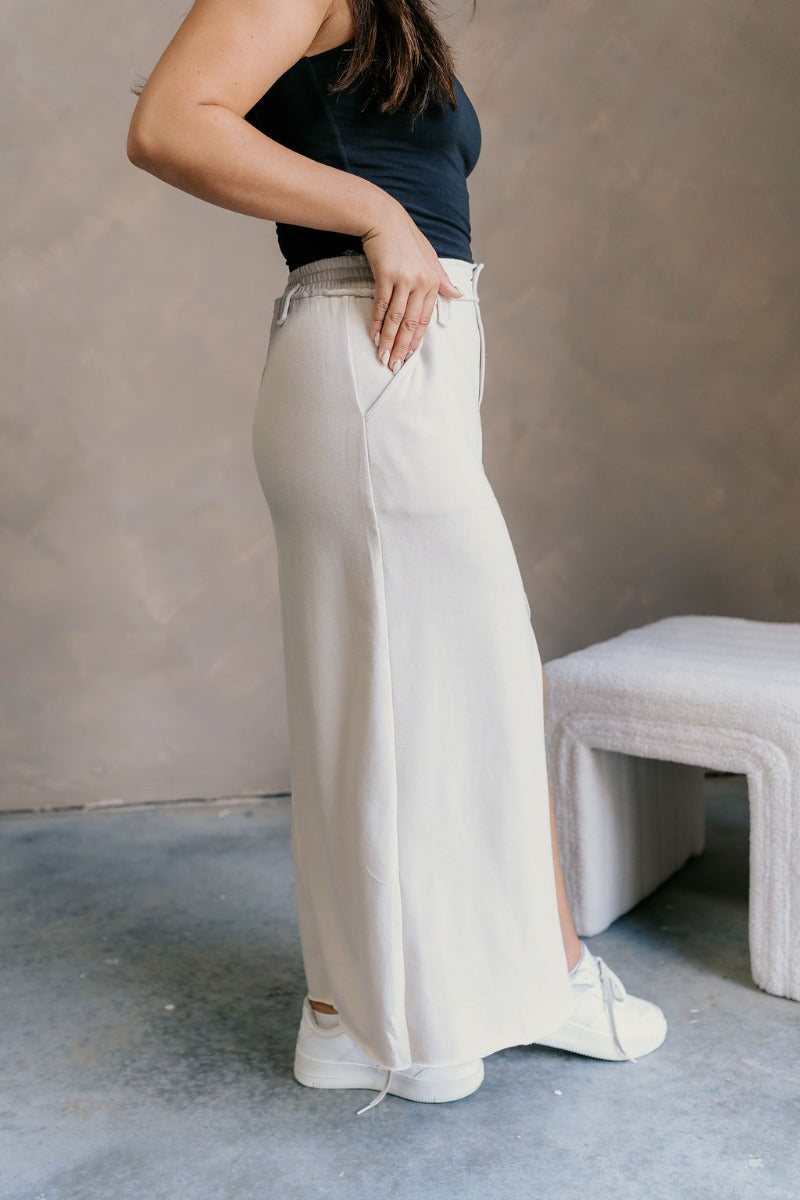 Side view of model wearing the Tori Cream Front Slit Midi Skirt which features cream fabric, a front slit, a tortoise button closure, a back elastic waistband, belt loops, front pockets, distressed details in the back, and a raw hem.