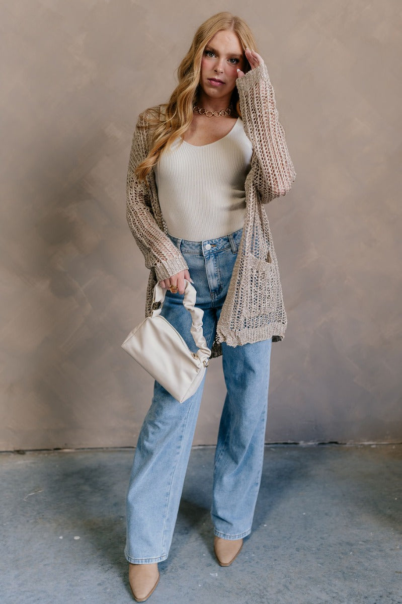 Full body view of model wearing the Remington Beige Crochet Knit Cardigan which features beige open knit fabric, a front opening with no closure, a thick hem, two front pockets, and long sleeves.