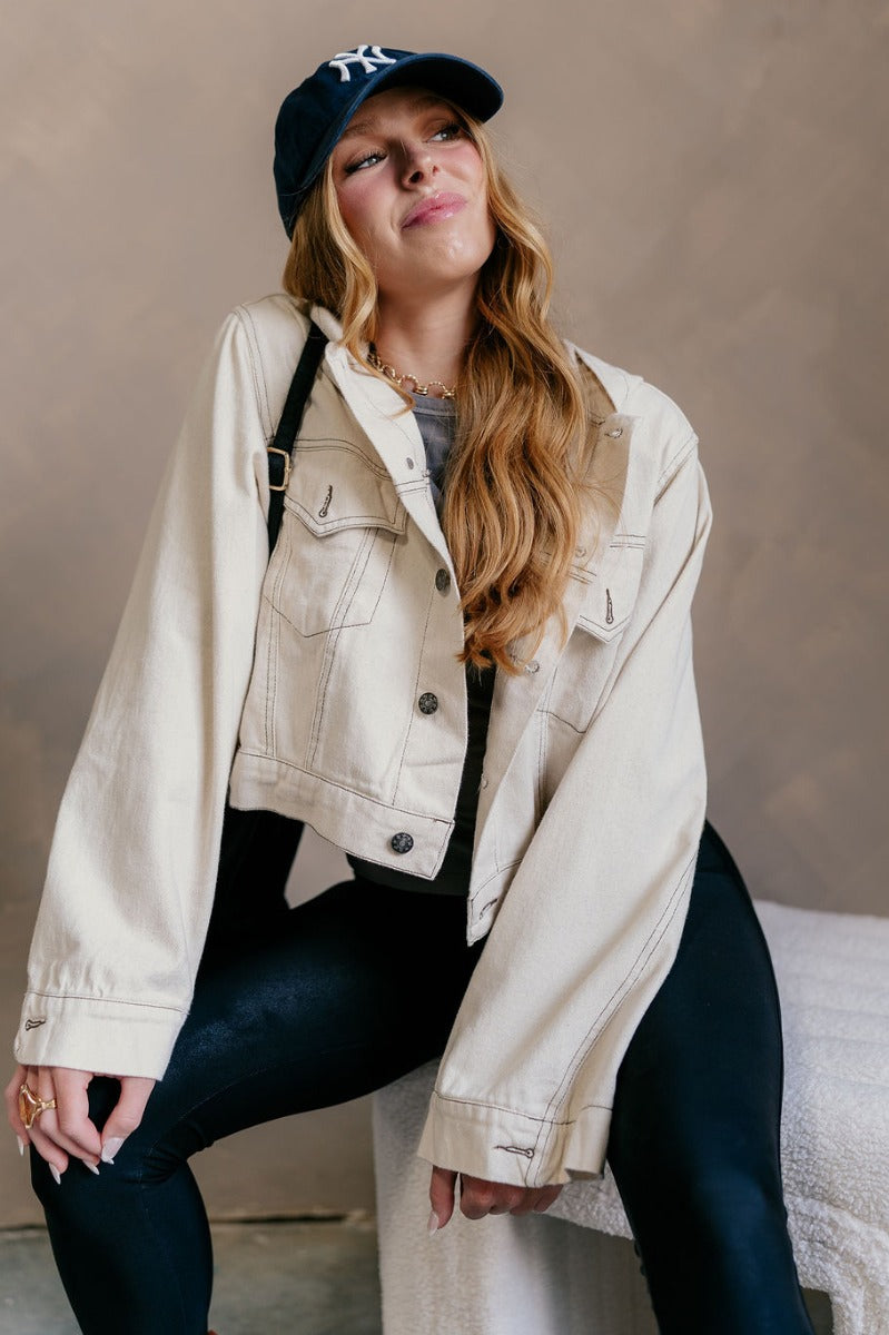 Front view of model wearing the Kennedy Cream Button-Up Hoodie Jacket which features oatmeal color cotton fabric, a cropped waist, star detail buttons, two front buttoned chest pockets, dark brown stitch detailing, a hood, and long flare sleeves with a bu