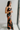 Side view of model wearing the Athena Black Multi Floral Slit Maxi Dress which features black satin fabric with a red, orange, and green rose floral pattern, maxi length, a slit on the side, a cowl neckline, adjustable spaghetti straps, and an open back w