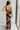 Back view of model wearing the Athena Black Multi Floral Slit Maxi Dress which features black satin fabric with a red, orange, and green rose floral pattern, maxi length, a slit on the side, a cowl neckline, adjustable spaghetti straps, and an open back w