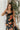 Front view of model wearing the Athena Black Multi Floral Slit Maxi Dress which features black satin fabric with a red, orange, and green rose floral pattern, maxi length, a slit on the side, a cowl neckline, adjustable spaghetti straps, and an open back 