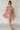 Full body view of model wearing the Iris Pink Multi Floral Mini Dress which features a hot pink, pink, grey, green, cream, mustard, mauve and light pink floral print, a v-neckline, tiered style, cream lining, pockets on each side, a smocked back, and shor