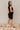 Back view of model wearing the Reese Black Sweater Sleeveless Dress features black knit fabric, mini length, belt loops with a tie around the waist, a high neckline, and a sleeveless design with padded shoulders.