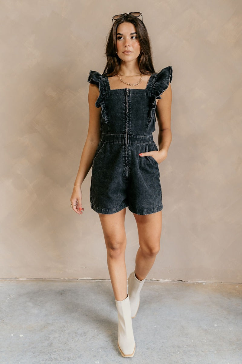 Full body view of model wearing the Avril Black Denim Ruffle Sleeveless Romper which features black washed denim fabric, pockets on each side, a front zip up, an elastic waistband, a square neckline, and ruffle straps.