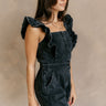 Front side view of model wearing the Avril Black Denim Ruffle Sleeveless Romper which features black washed denim fabric, pockets on each side, a front zip up, an elastic waistband, a square neckline, and ruffle straps.
