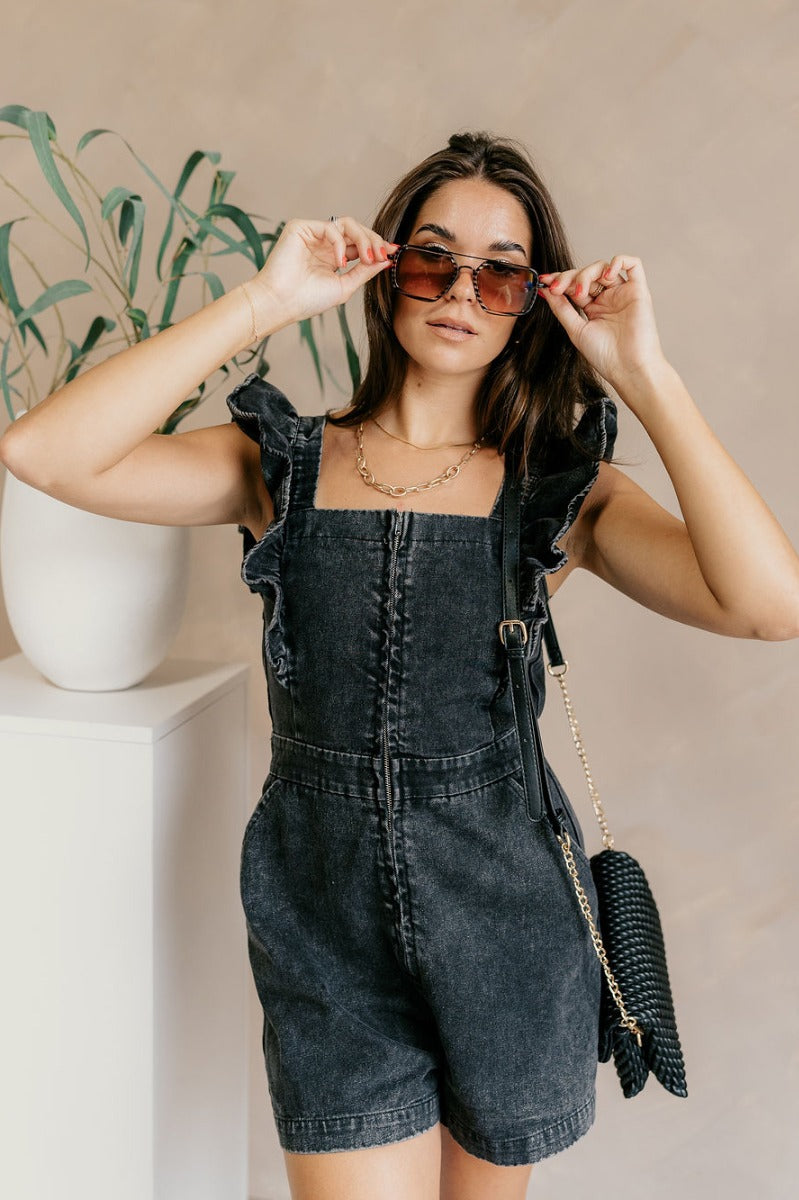 Front view of model wearing the Avril Black Denim Ruffle Sleeveless Romper which features black washed denim fabric, pockets on each side, a front zip up, an elastic waistband, a square neckline, and ruffle straps.