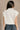 Back view of model wearing the Blakely Cream High Neck Sleeveless Sweater which features cream cable knit fabric, a thick hem, a high neckline and a sleeveless design.