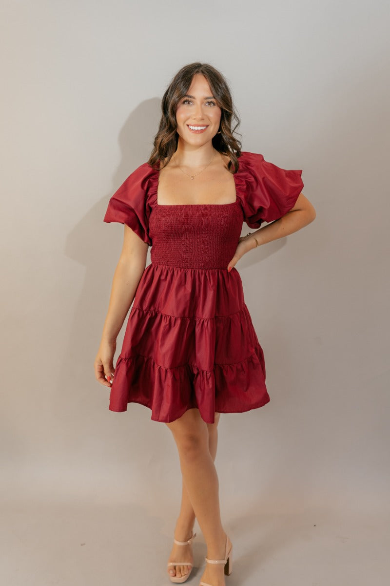 Full body front view of model wearing the Willow Burgundy Puff Sleeve Dress that has burgundy fabric, mini length, a tiered skirt, smocked chest, a square neck and short puff sleeves