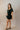 Full body side view of model wearing the Eliana Black Tiered Short Sleeve Romper that has black lightweight fabric, a ruffle-tiered skirt, black shorts lining, an elastic waistband, a sweetheart neckline and short puff sleeves.