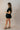 Full body side view of model wearing the Eliana Black Tiered Short Sleeve Romper that has black lightweight fabric, a ruffle-tiered skirt, black shorts lining, an elastic waistband, a sweetheart neckline and short puff sleeves.