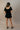 Full body back view of model wearing the Eliana Black Tiered Short Sleeve Romper that has black lightweight fabric, a ruffle-tiered skirt, black shorts lining, an elastic waistband, a sweetheart neckline and short puff sleeves.