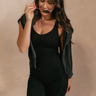 Front view of model wearing the Amara Black Athletic Onesie Bodysuit that has black atheisure fabric, side pockets, built-in-padding, monochrome stitching, and thick straps. Sweater is draped on model's shoulders.