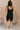 Full body back view of model wearing the Amara Black Athletic Onesie Bodysuit that has black atheisure fabric, side pockets, built-in-padding, monochrome stitching, and thick straps. Sweater is draped on model's shoulders.