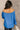 Back view of model wearing the Audrey Blue Satin Adjustable Neckline Long Sleeve Top which features blue satin fabric, a round neckline with drawstring tie, and long balloon sleeves with elastic wrists.