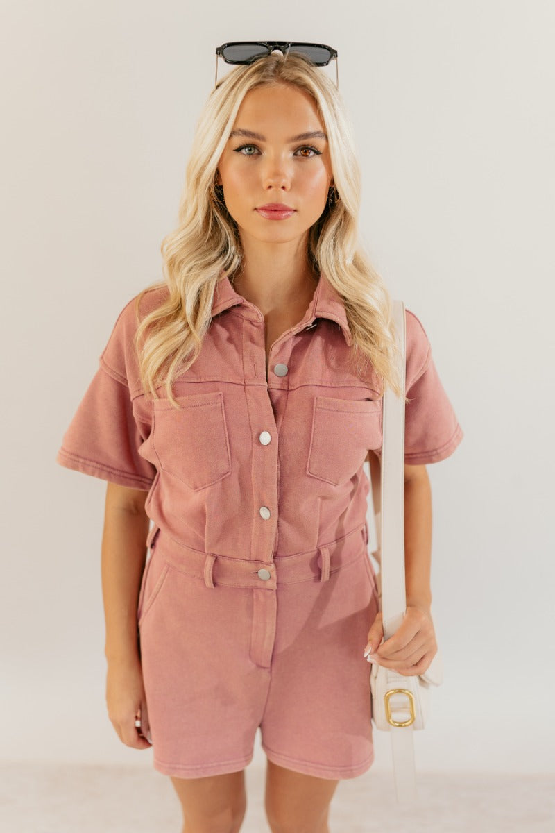 front view of model wearing the Sadie Pink Knit Short Sleeve Romper that has washed pink knit fabric, pockets, a front zipper, a collar, belt loops, an elastic waistband, and short sleeves.