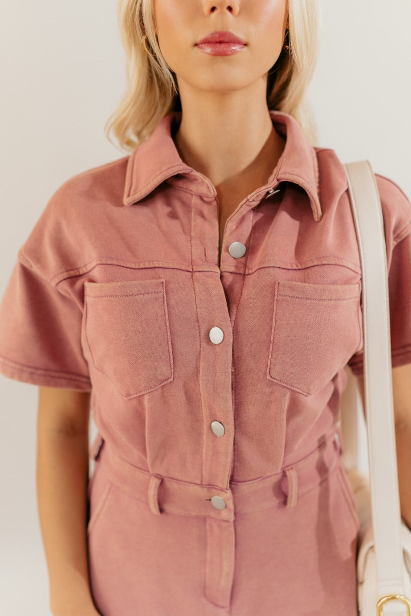 Close upper front view of model wearing the Sadie Pink Knit Short Sleeve Romper that has washed pink knit fabric, pockets, a front zipper, a collar, belt loops, an elastic waistband, and short sleeves.