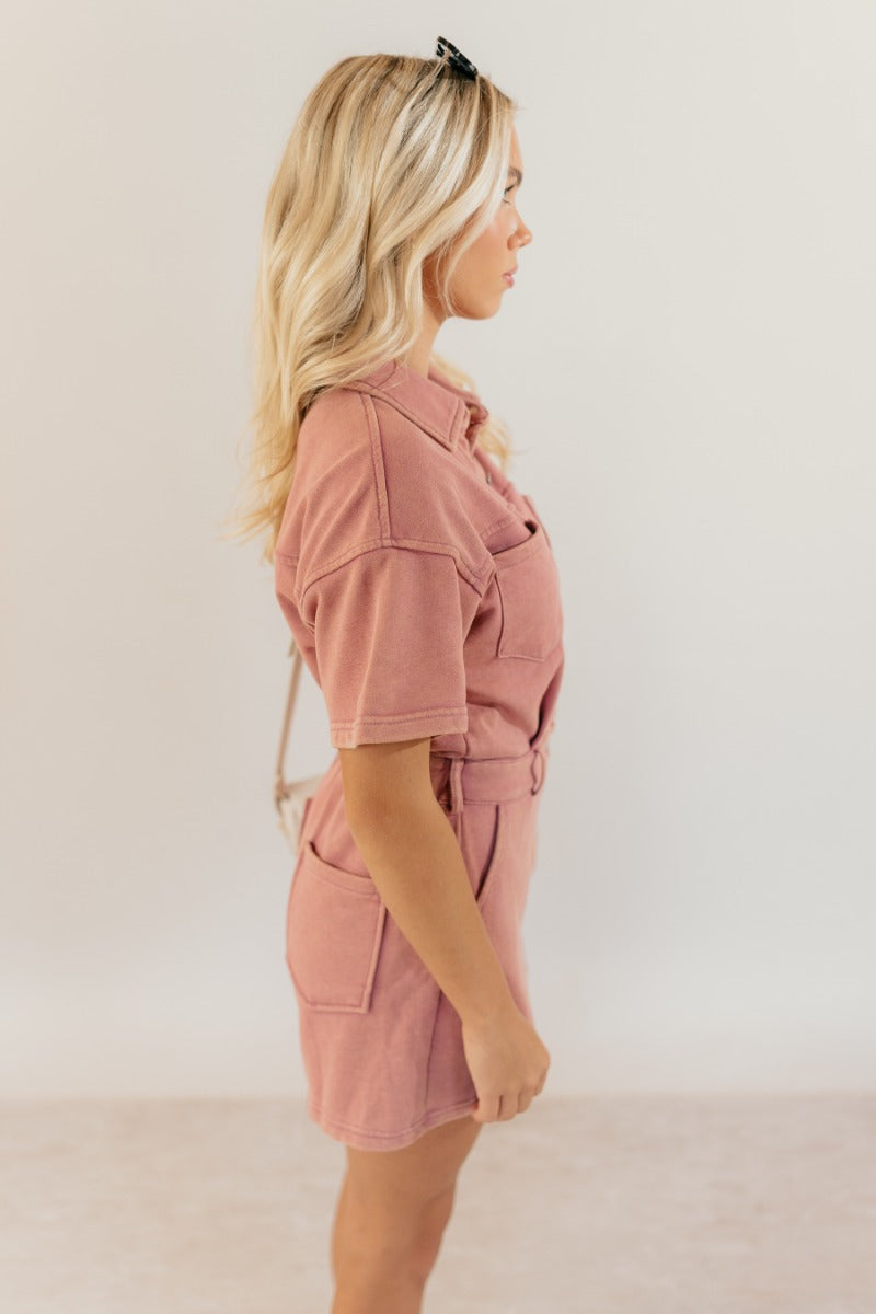 Side view of model wearing the Sadie Pink Knit Short Sleeve Romper that has washed pink knit fabric, pockets, a front zipper, a collar, belt loops, an elastic waistband, and short sleeves.