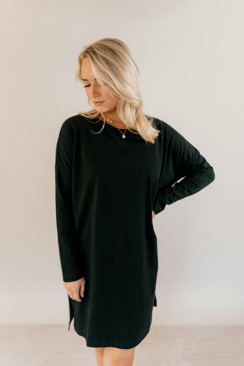 front view of model wearing the Sara Black Long Sleeve T-shirt Dress that has black cotton fabric, mini length, slits on each side, a round neckline, dropped shoulders, and long sleeves.