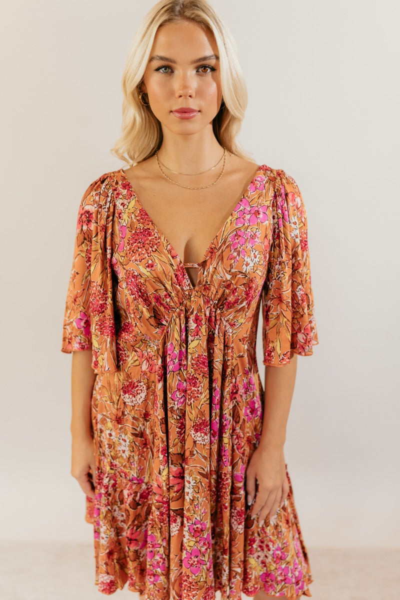 front view of model wearing the Rachel Rust Floral Short Sleeve Mini Dress that has rust fabric with a floral pattern, mini length, flared skirt, a plunge neck, an open back with a tie , and short flare sleeves.