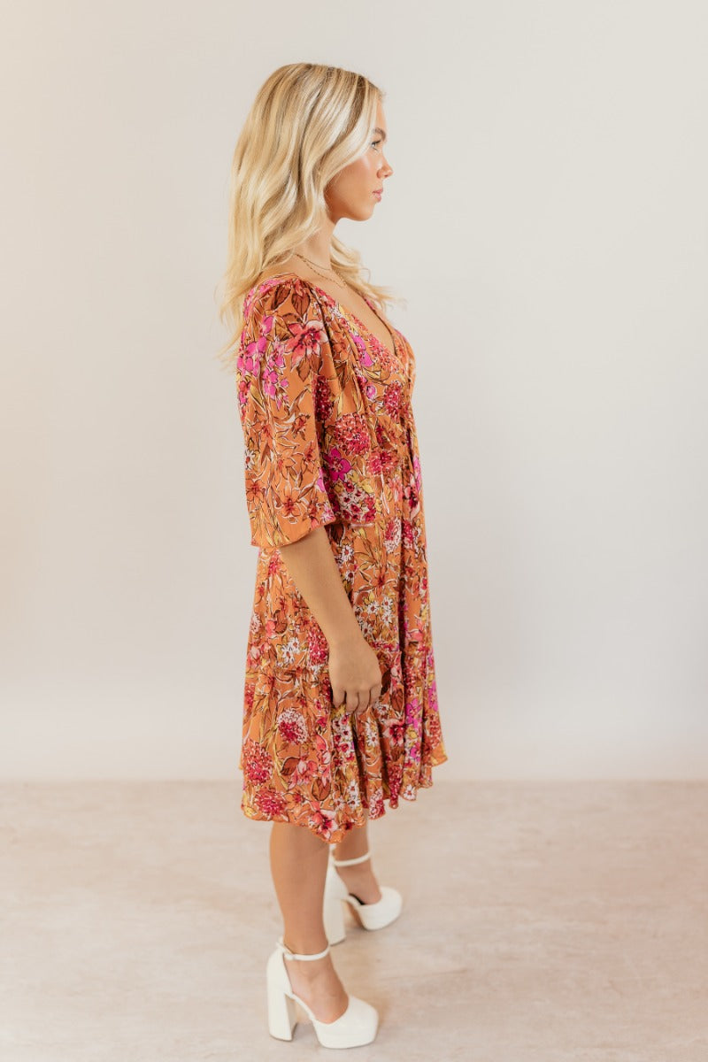 Full body side view of model wearing the Rachel Rust Floral Short Sleeve Mini Dress that has rust fabric with a floral pattern, mini length, flared skirt, a plunge neck, an open back with a tie , and short flare sleeves.