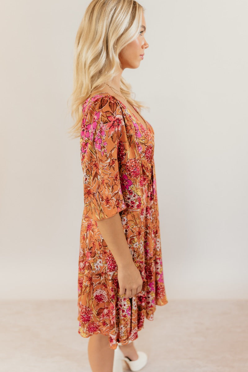 Side view of model wearing the Rachel Rust Floral Short Sleeve Mini Dress that has rust fabric with a floral pattern, mini length, flared skirt, a plunge neck, an open back with a tie , and short flare sleeves.