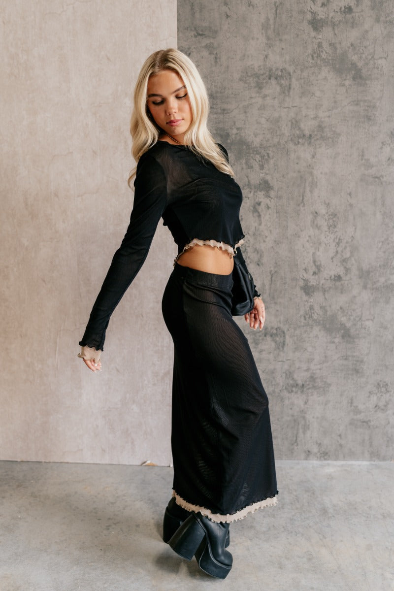 Full body side view of model wearing the Kylie Black & Taupe Mesh Midi Skirt that has black mesh fabric, taupe mesh lining, an elastic waist, a slit on the side, and lettuce hem details.