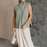 Full body front view of model wearing the Wren Sage Sleeveless Turtleneck Sweater that features sage green knit fabric, a high-low hem, slits on each side, a turtleneck neckline and a sleeveless design.