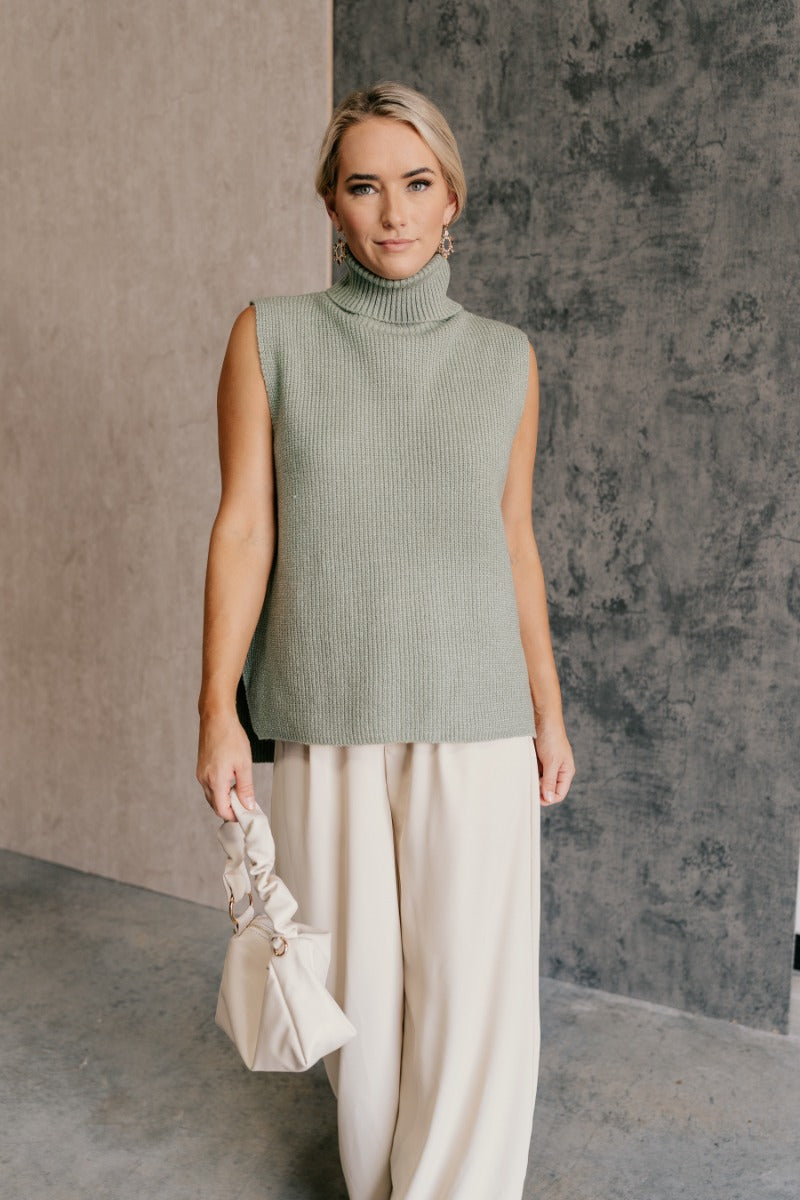 front view of model wearing the Wren Sage Sleeveless Turtleneck Sweater that features sage green knit fabric, a high-low hem, slits on each side, a turtleneck neckline and a sleeveless design.