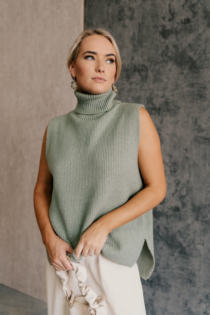 front view of model wearing the Wren Sage Sleeveless Turtleneck Sweater that features sage green knit fabric, a high-low hem, slits on each side, a turtleneck neckline and a sleeveless design.