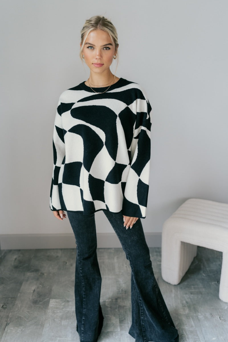 Full front view of model wearing the Lennon Black & White Geometric Sweater that has black and white knit fabric with a geometric pattern, a round neckline, dropped shoulders, and long flare sleeves.