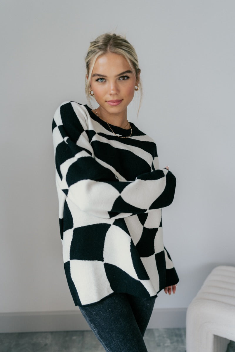 Front view of model wearing the Lennon Black & White Geometric Sweater that has black and white knit fabric with a geometric pattern, a round neckline, dropped shoulders, and long flare sleeves.