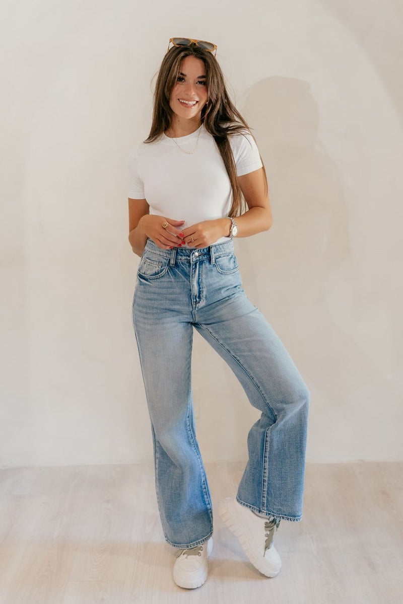 Full body front view of model wearing the The Rooted Denim: Camila Wide Leg Jeans that have light medium denim fabric, a front zipper with a button closure, belt loops, pockets and wide legs.