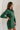 Side view of model wearing the Gianna Dark Green Button Front Dress that has dark green fabric, white stitch detailing, front button closures, a collared neck, chest pockets, and long sleeves.