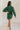 Full body back view of model wearing the Gianna Dark Green Button Front Dress that has dark green fabric, white stitch detailing, front button closures, a collared neck, chest pockets, and long sleeves.