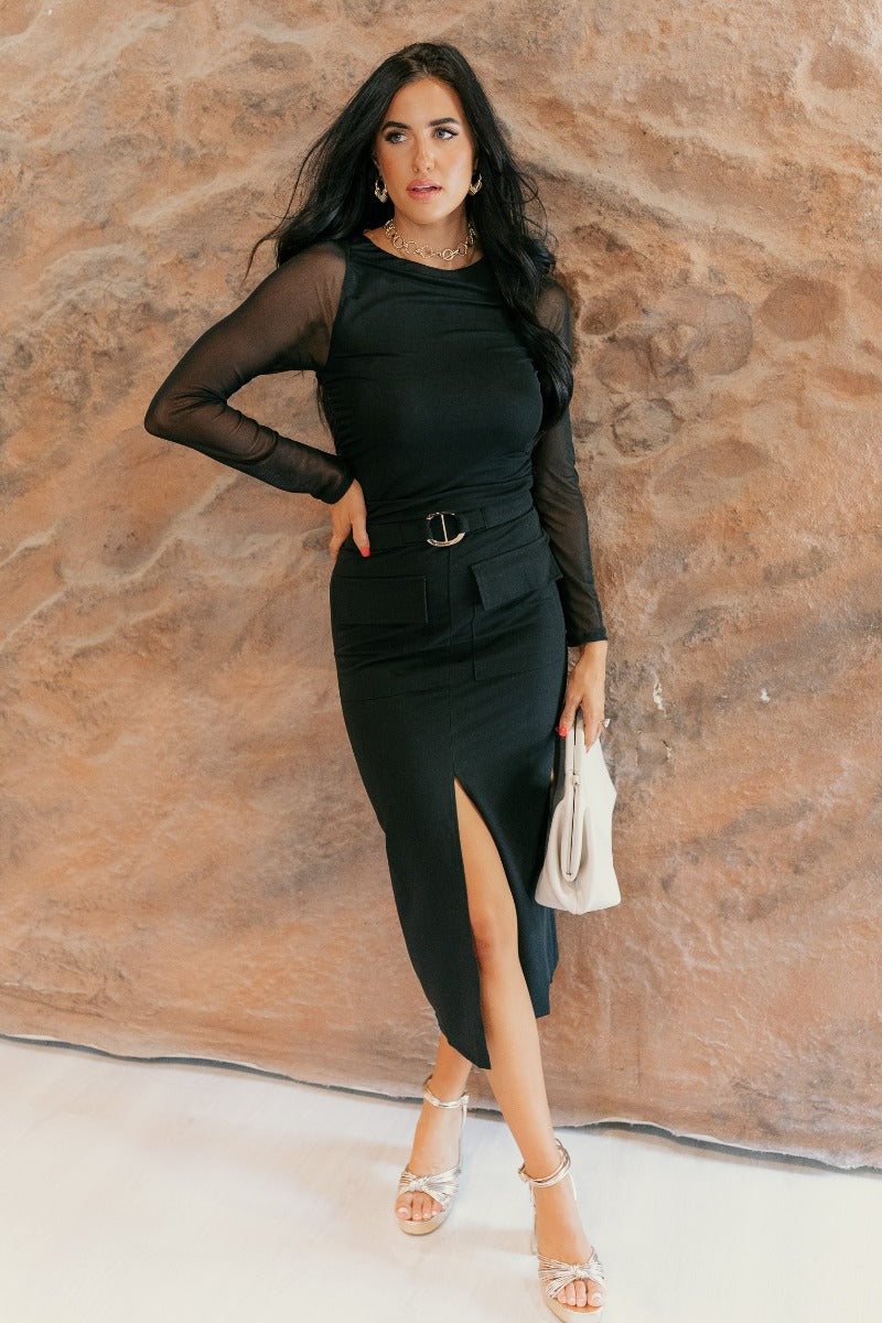 Full body front view of model wearing the Hailey Belted Black Front-Slit Midi Skirt that has black lightweight fabric, cargo pockets, a side zipper with a hook, a shiny gold hoop belt detail, midi length, and a front slit.