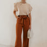 Full body front view of model wearing the Audrey Rust Wide Leg Pants that have rust lightweight fabric, two front pockets, a front zipper with a hook closure, a tie closure with matte gold adjustable hoops, and wide legs.