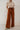 front view of model wearing the Audrey Rust Wide Leg Pants that have rust lightweight fabric, two front pockets, a front zipper with a hook closure, a tie closure with matte gold adjustable hoops, and wide legs.