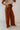 Side view of model wearing the Audrey Rust Wide Leg Pants that have rust lightweight fabric, two front pockets, a front zipper with a hook closure, a tie closure with matte gold adjustable hoops, and wide legs.