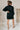 Full body back view of model wearing the Laura Black Sweater Belted Dress which features black knit fabric, mini length, a monochromatic adjustable belt at the waistline, a high neckline and long balloon sleeves.