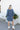 Full body back view of model wearing the Away We Go Dress, that has blue washed fabric, a two-tiered babydoll style, a v neckline, and 3/4 balloon sleeves with elastic wrists.