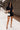 Full body front view of model wearing the Running The Show Denim Romper, which features black washed denim, a surplice neck that snaps with a collar, half-length sleeves, a front zipper, pockets, pleating on the legs, and a 3" inseam