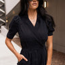 Front view of model wearing the Running The Show Denim Romper, which features black washed denim, a surplice neck that snaps with a collar, half-length sleeves, a front zipper, pockets, pleating on the legs, and a 3" inseam