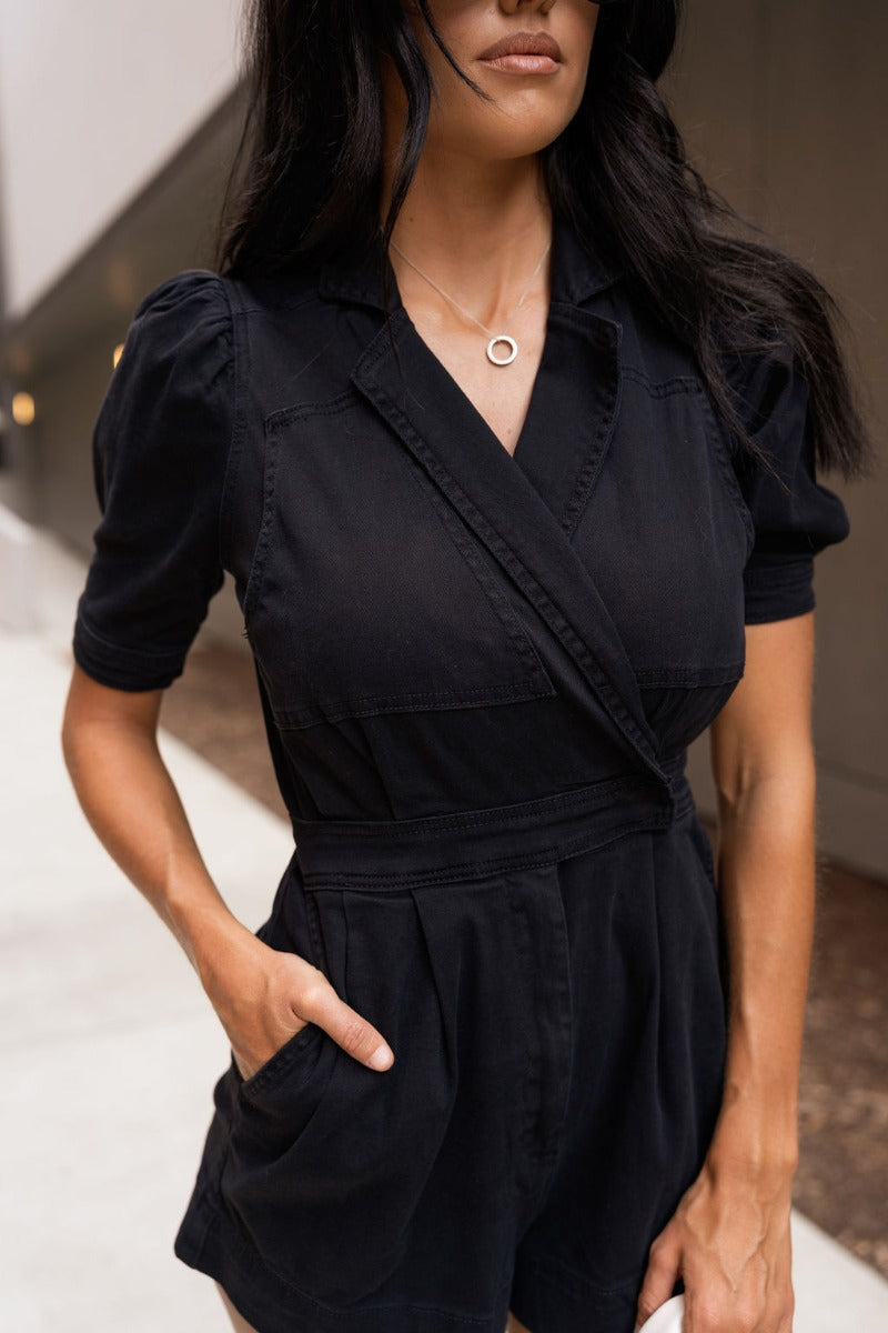 Close-up front view of model wearing the Running The Show Denim Romper, which features black washed denim, a surplice neck that snaps with a collar, half-length sleeves, a front zipper, pockets, pleating on the legs, and a 3" inseam