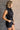 Side view of model wearing the Emily Washed Black Denim Sleeveless Romper which features washed black denim fabric, two front pockets, silver button details, button up front closure, two front chest buttoned pockets, two back pockets, collared neckline an