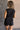 Back view of model wearing the Emily Washed Black Denim Sleeveless Romper which features washed black denim fabric, two front pockets, silver button details, button up front closure, two front chest buttoned pockets, two back pockets, collared neckline an