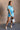 Full body view of model wearing the Delilah Washed Sky Blue Short Sleeve Romper which features washed light blue tencel fabric, two front slit pockets, an elastic waistband with a drawstring tie, a right front chest pocket, tortoise button closures, a col