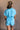 Back view of model wearing the Delilah Washed Sky Blue Short Sleeve Romper which features washed light blue tencel fabric, two front slit pockets, an elastic waistband with a drawstring tie, a right front chest pocket, tortoise button closures, a collared